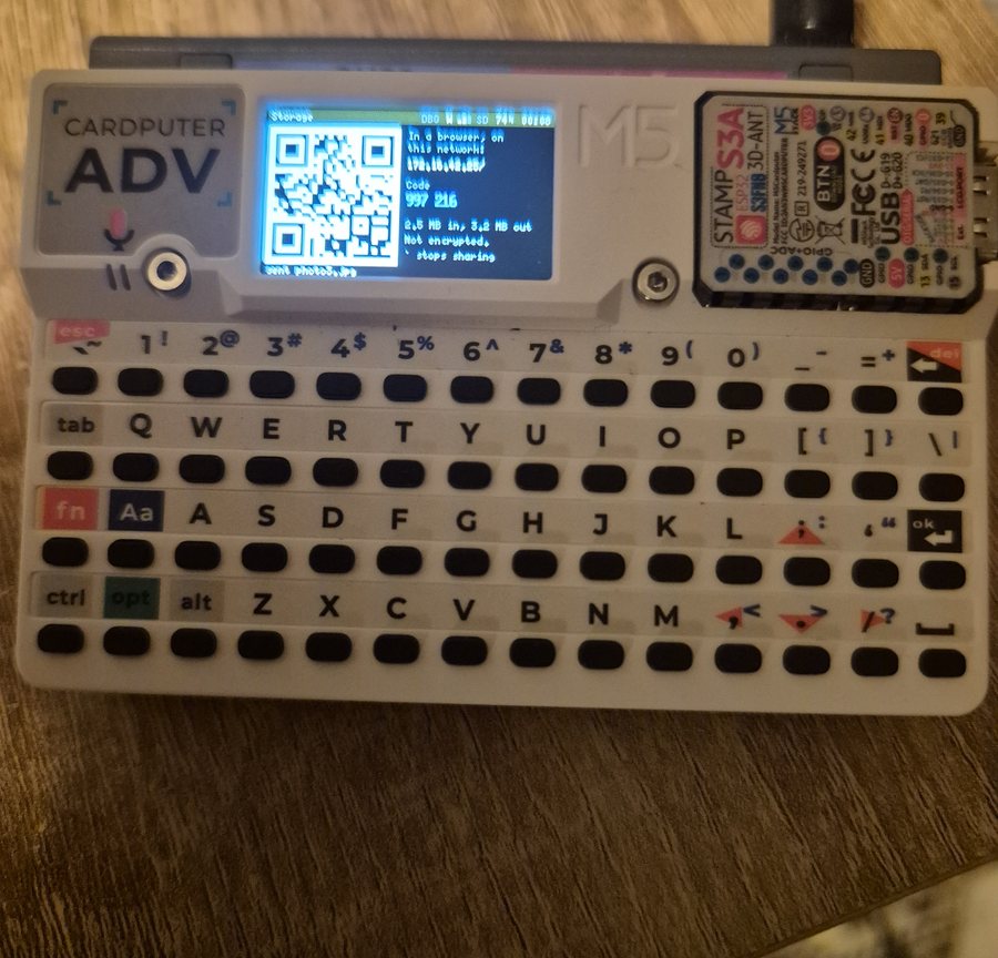 A photograph of the Cardputer ADV on a wooden table. Its small screen shows the QR code, the address, the code 997 216, and 2.5 MB in, 3.2 MB out. Below the screen, the whole keyboard.
